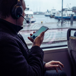 Torso og hænder på en pendlende person i en nordisk sporvogn, med headset og smartphone tæt på munden; dybe indigo‑skygger og cyan signalaccents antyder realtids stemme‑streaming uden synlig tekst.