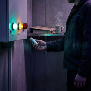 Operationsniche i en dansk SMV: en tekniker (kun torso) holder en forsejlet checkpoint‑kapsel op mod en vægmonteret statuslampe, amber‑lyset pulserer. Stemning: kølig nordisk skumring, indikation af gating, risiko og beslutningsøjeblik.