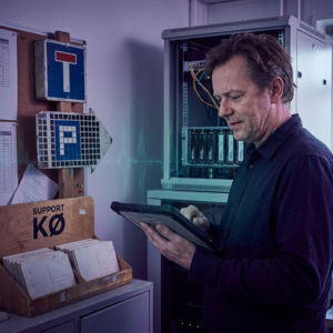 Teknisk leder i et lille operationsrum overvejer et arkitekturvalg; NVMe‑enheder og en åben rackhylde synlig, supportkø som fysiske kort, to rute‑skilte som metafor for tekst vs pixels. Nordisk, indigo/purpur tone med cyan‑grønne accenter.