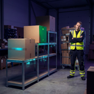 Dansk dispatch‑hal med cyan‑markeret gulvspor og pakker; en operationsleder i arbejdsjakke observerer et hold i flowet. Indigo/teal tone, ingen tekst.