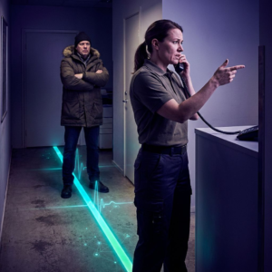 Medarbejder i et lille nordisk serviceområde taler i telefon mens en klient venter ved en cyan/gulvstribe; dybe indigo‑skygger og cyan/green lysaccents skaber en filmisk, beslutningsfokuseret stemning.