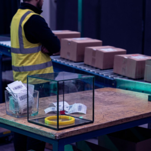 Nordisk operationsmiljø: et gennemsigtigt test‑'terrarium' på vogn i forgrunden, dispatch‑kø med pakker i baggrunden og en bekymret operationsleder i periferien — blå‑lilla stemning med cyan/green accenter.