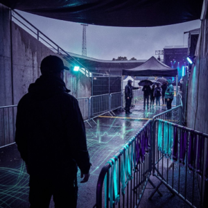 Backstage ved et nordisk arena‑indgang: operationsleder i silhuet, to farvekodede armbånd på barrierer, personale omdirigerer en våd kø, indigo og cyan accentlys, subtil data‑pulse overlay.