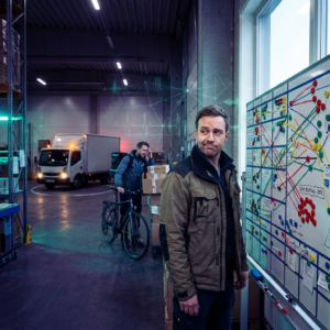 Nordisk operationsleder i et lille lager ser på farvekodede rute‑markører på gulvet; cyan/grønne accenter og dybe indigo skygger antyder teknisk routing‑beslutning uden læsbar tekst.