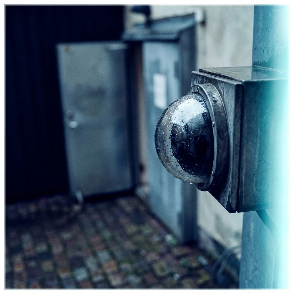 Sikkerhedskamera monteret i en baggård med indigo lys og cyan refleks, servicedør let åben i baggrunden — metafor for overvågnings‑og telemetri‑behov.