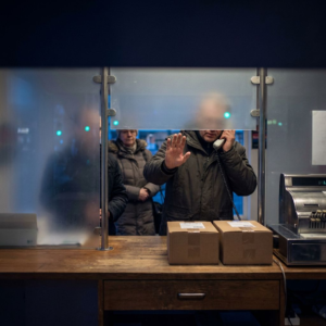En service-disk i en lille dansk pakkeshop, hvor en hånd stopper en lille pakke foran en halvt nedrullet luge, mens en kort kø venter i baggrunden