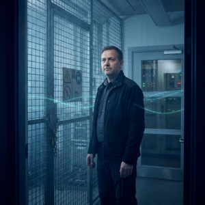 Teknisk leder stående foran en låst testsluse i et nordisk operationsrum, dybe indigo‑skygger og cyan/grønne signalreflekser — symbol på valg mellem åben benchmark‑innovation og stram driftssikring.