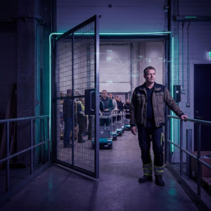 Dansk operationsleder ved en åben sikkerhedssluse i en lille fabrikshal, med ventende produkter bag slusen og indigo/cyan lysning, antydning af en diskret datapuls over slusen.