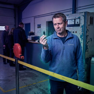 Operationsleder ved en kømærket servicepassage i en dansk virksomhed, ser over på en slidt maskin‑stub mens han holder en lille test‑node; indigo/lilla lys og cyan/grønne accenter antyder data og moderniseringsvalg.