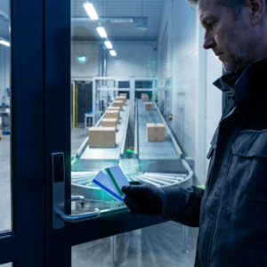 Nordisk indgangssluse til et produktionsområde, en mellemleder holder to farvekodede adgangskort foran en glasdør, mens et transportbånd bag glasset deler sig i to ruter.
