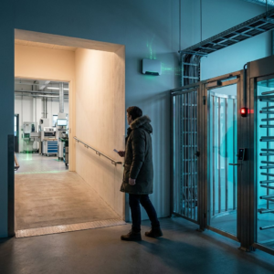 En driftansvarlig står ved en dansk adgangspassage med to ruter – én åben og én med turnstile – og tøver et øjeblik i køligt nordisk morgenlys uden læsbar skiltning.
