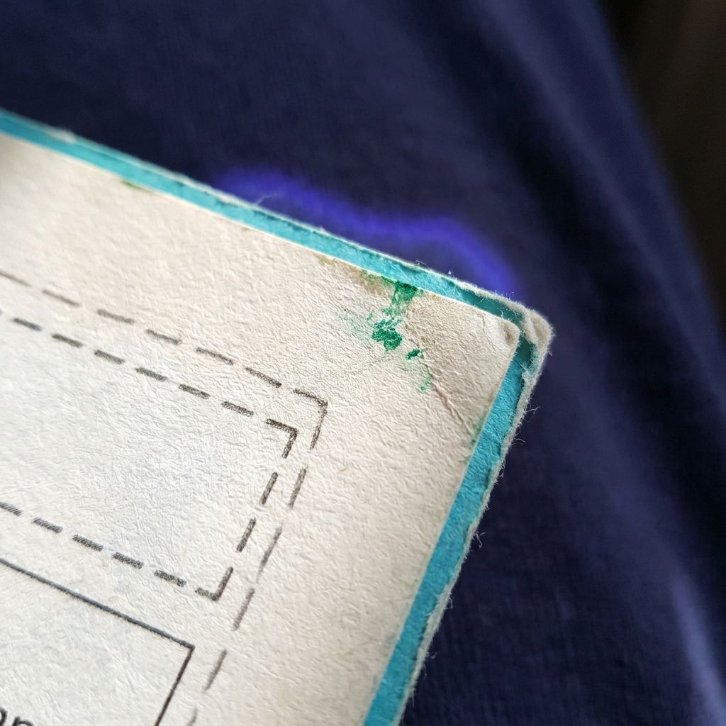 Tæt dokumentarisk makro af en papirkant med et rektangulært mærke som symbol på en bounding box; fibrene og let slid synlige, dyb indigo baggrund.