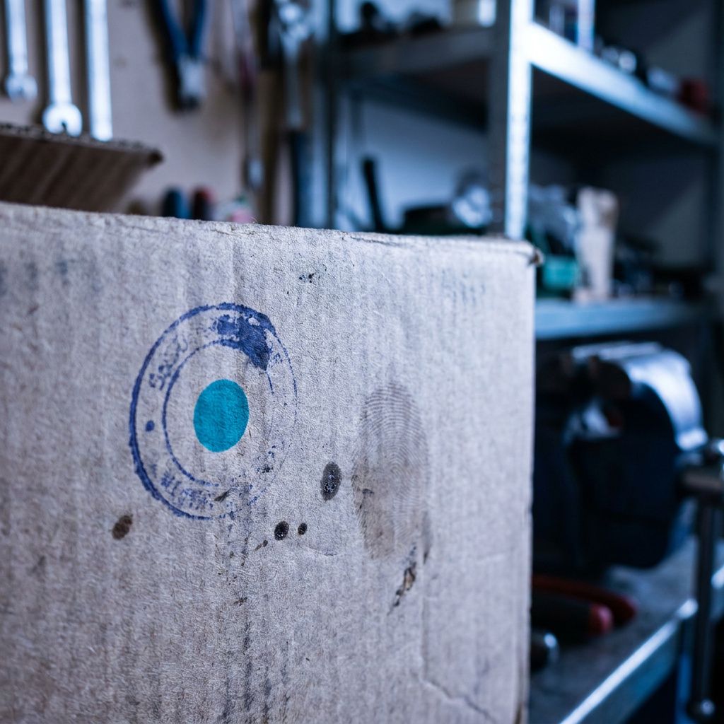 Makrodetalje af en stempelmærket mærkat på en kasse: slidte kanter, en indigo streg, et cyan punkt, fingeraftryk, spor af transportstøj.