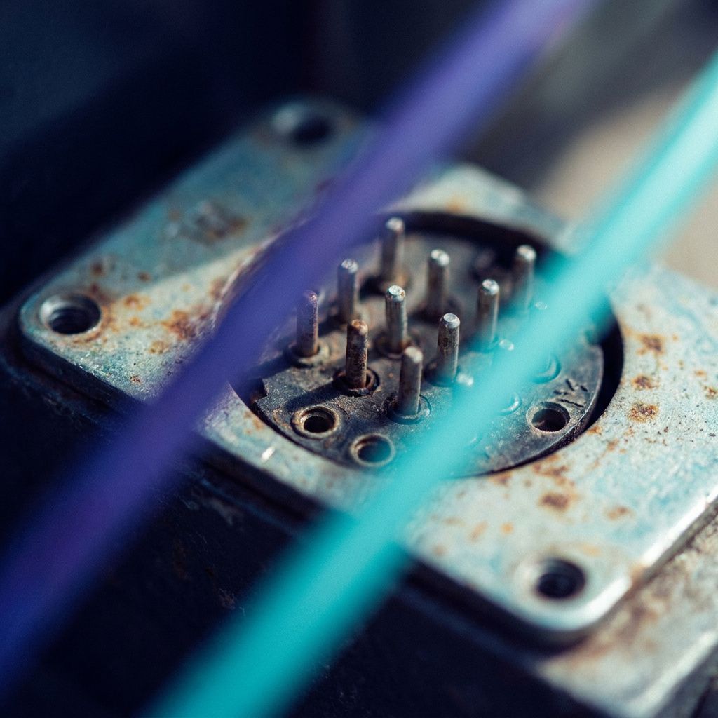Makrofoto af en slidt metalplade på en testboks med indigo og cyan lysreflektioner, kort dybdeskarphed, subtil pulserende glød i baggrunden.