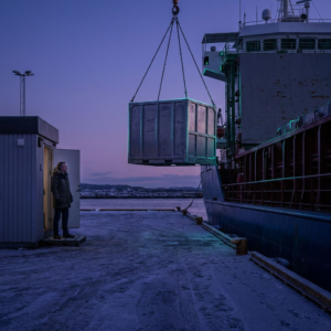 En nordisk IT‑chef ved dørtærsklen til et aflåst teknisk rum ser på en forsejlet transportkasse; dybe lilla skygger, cyan/grønne signalreflekser, subtil datapuls‑overlay.