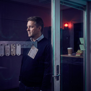Nordisk SMV‑leder foran en supporthub, beslutsom, med indigo og cyan accent – visualiserer valg mellem AI‑modeller målt på cost‑per‑outcome.