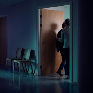 Diskret triagescene i nordisk klinikkorridor, hvor en medarbejder guider en borger ind i et privat rum, mens tomme stole antyder ventekø