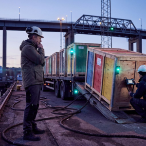 En nordisk teknisk leder i profil på kajen ved en lille havn, ser på en række farvekodede kasser på en trailer der forlader stedet — billedet symboliserer netværks‑ og leveringskøen mellem rollout og træning i MoE‑RL